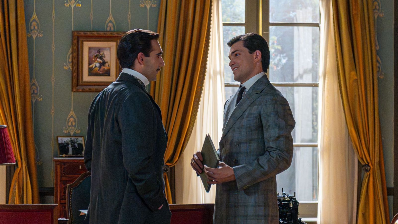 Dos hombres conversan en una estancia señorial decorada con papel tapiz verde y cortinas doradas, vistiendo elegante ropa de principios del siglo XX.