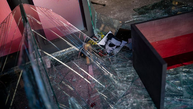 Plano picado de los destrozos en un escaparate comercial con cristales de seguridad fracturados, un expositor volcado y restos de publicidad en el suelo.