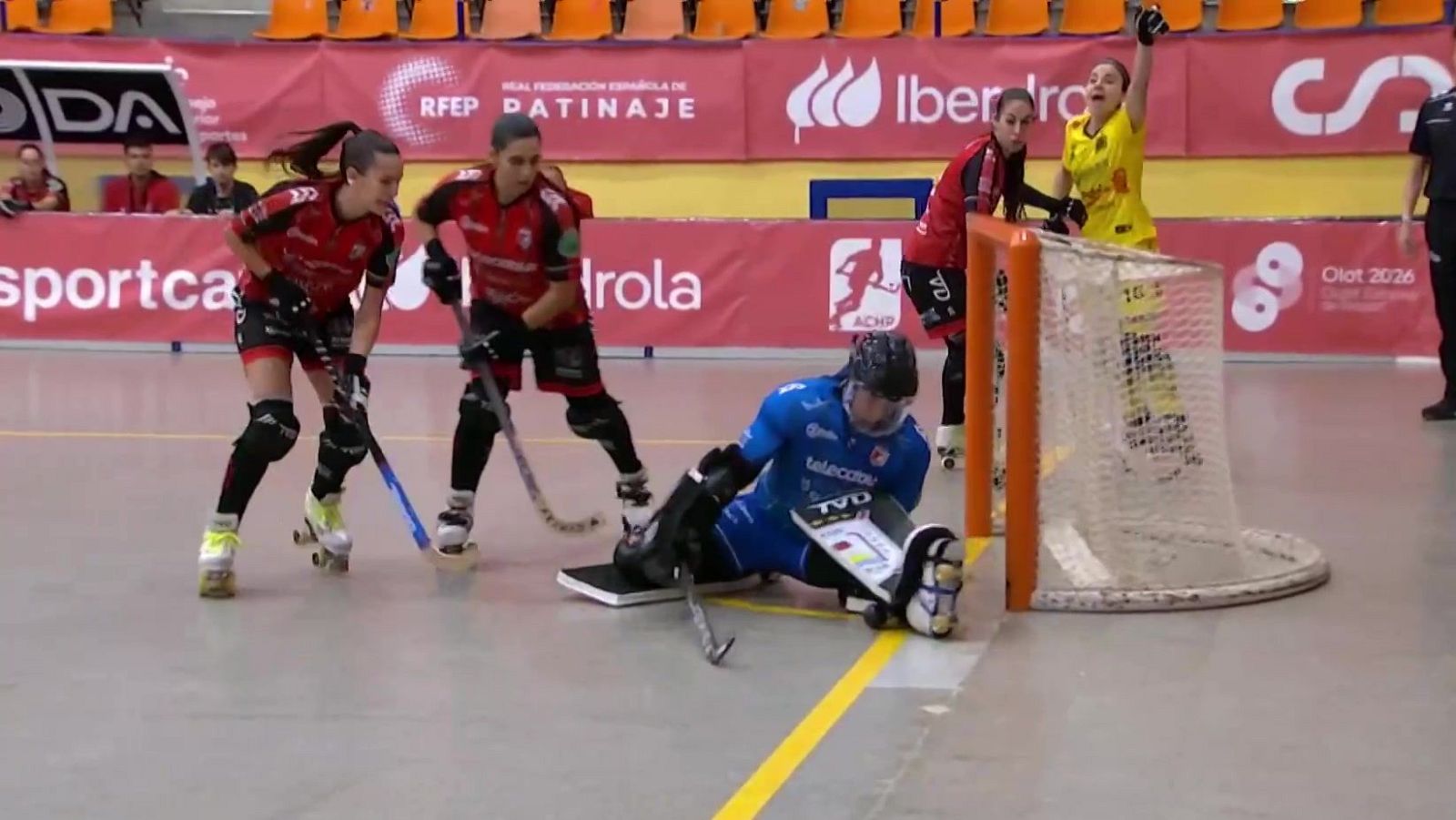 Partido de hockey sobre patines femenino con portera en el suelo junto a la portería, jugadoras con uniforme rojo y una rival celebrando.