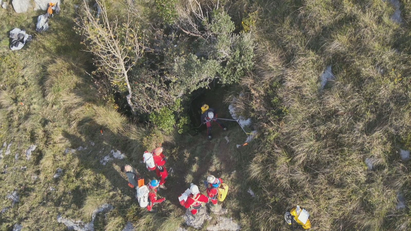 Plano cenital de un equipo de emergencia con monos rojos y cascos preparando un descenso a una sima natural en terreno montañoso.