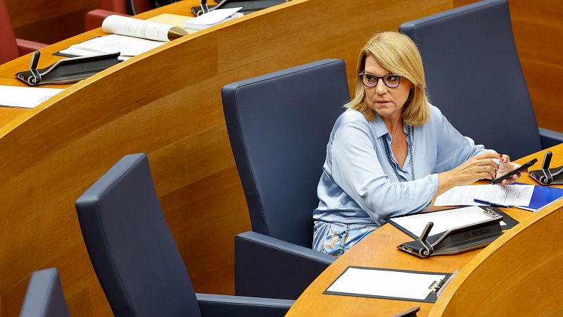 Mujer rubia con gafas y blusa azul celeste sentada en un escaño parlamentario, sosteniendo un teléfono móvil con documentos sobre la mesa.
