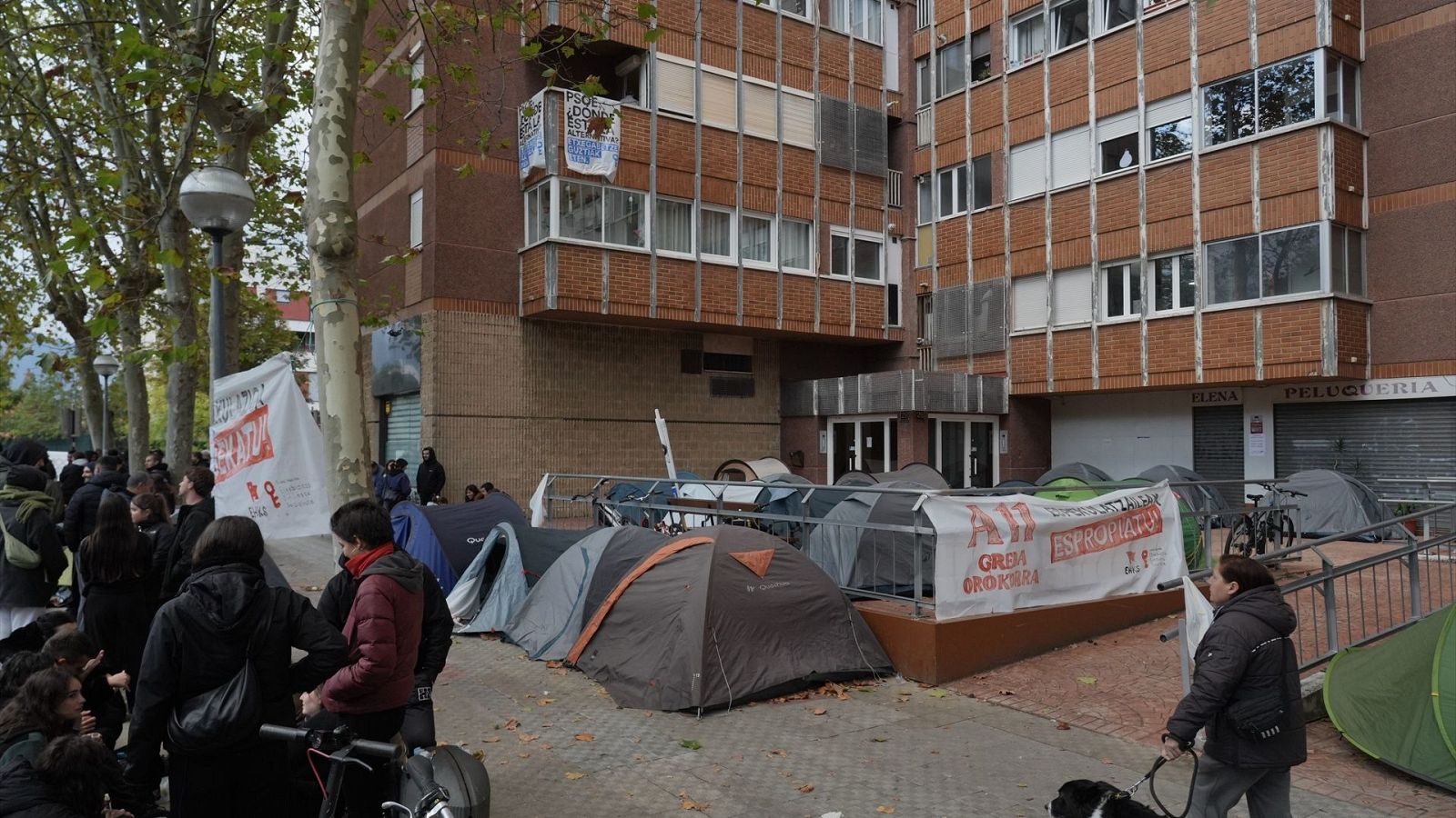 Protesta con tiendas de campaña y pancartas frente a un edificio residencial, con manifestantes en la acera y árboles otoñales.