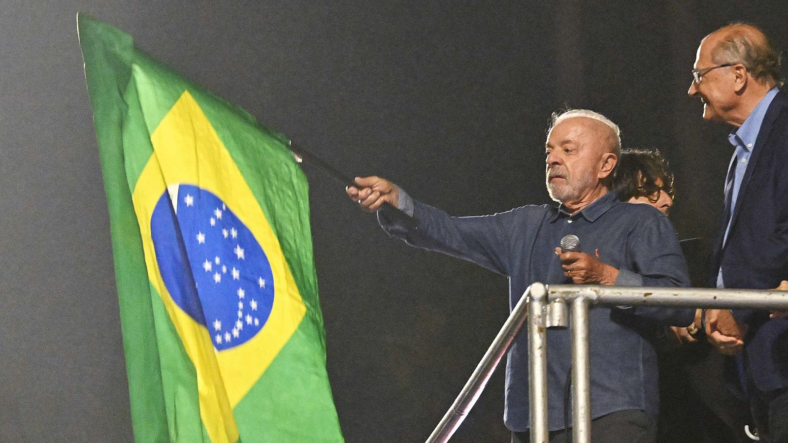 Fotografía nocturna de Luiz Inácio Lula da Silva sobre una plataforma ondeando una gran bandera de Brasil junto a Geraldo Alckmin.