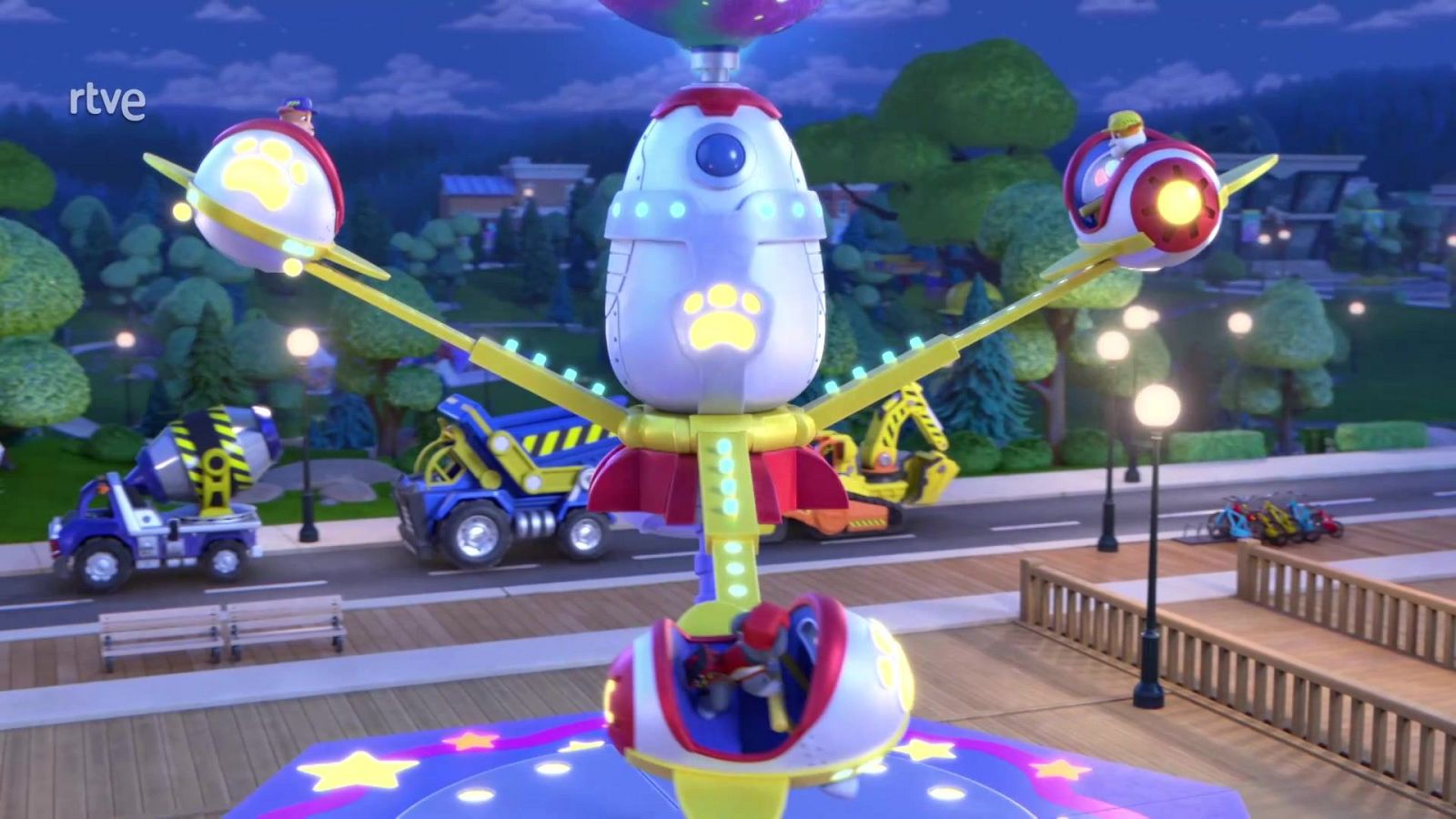 Atracción de feria infantil iluminada con cápsulas espaciales y cachorros animados, junto a vehículos de construcción al atardecer.