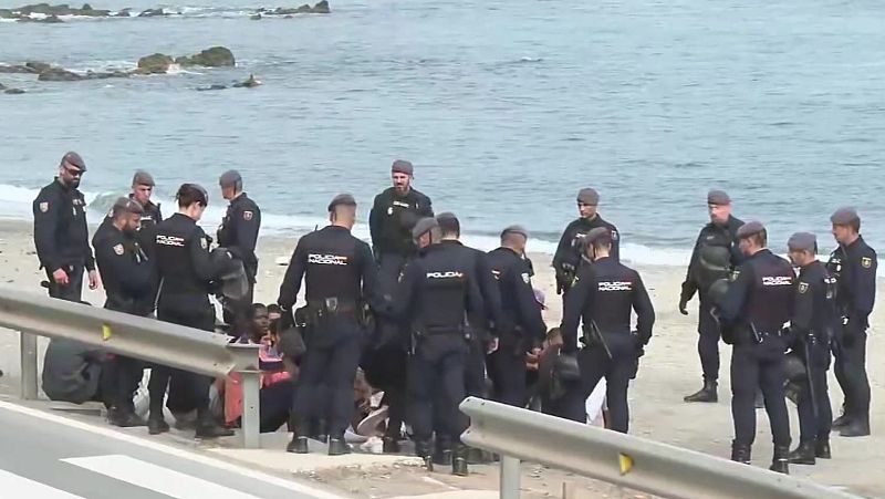 Agentes policiales uniformados custodian a varias personas sentadas en la orilla de una playa junto a una carretera, bajo un cielo nublado.