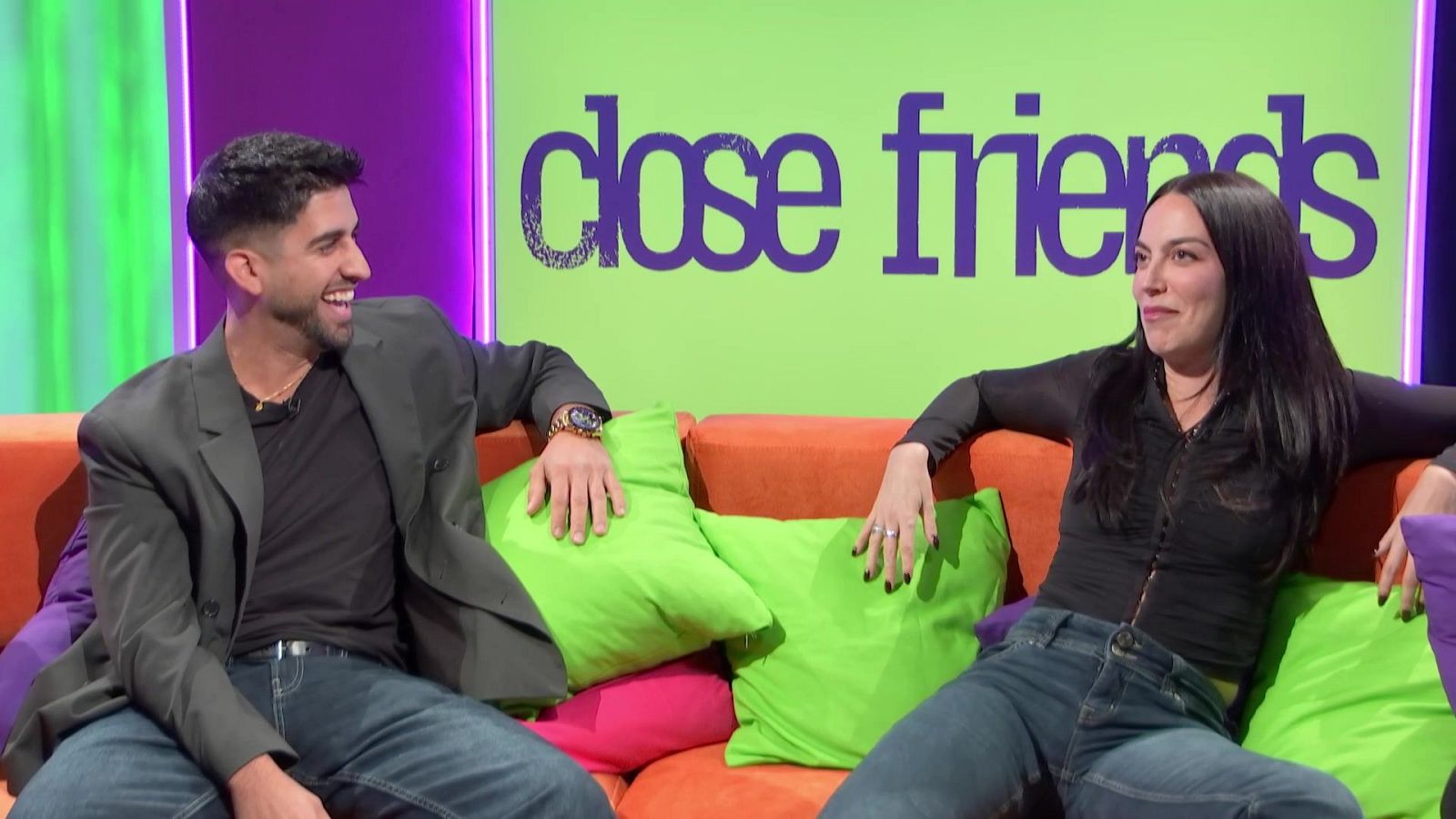 Dante Caro y María Valero charlan sonrientes sentados en un sofá naranja en el plató del programa Close Friends, decorado con paneles de colores y luces LED.