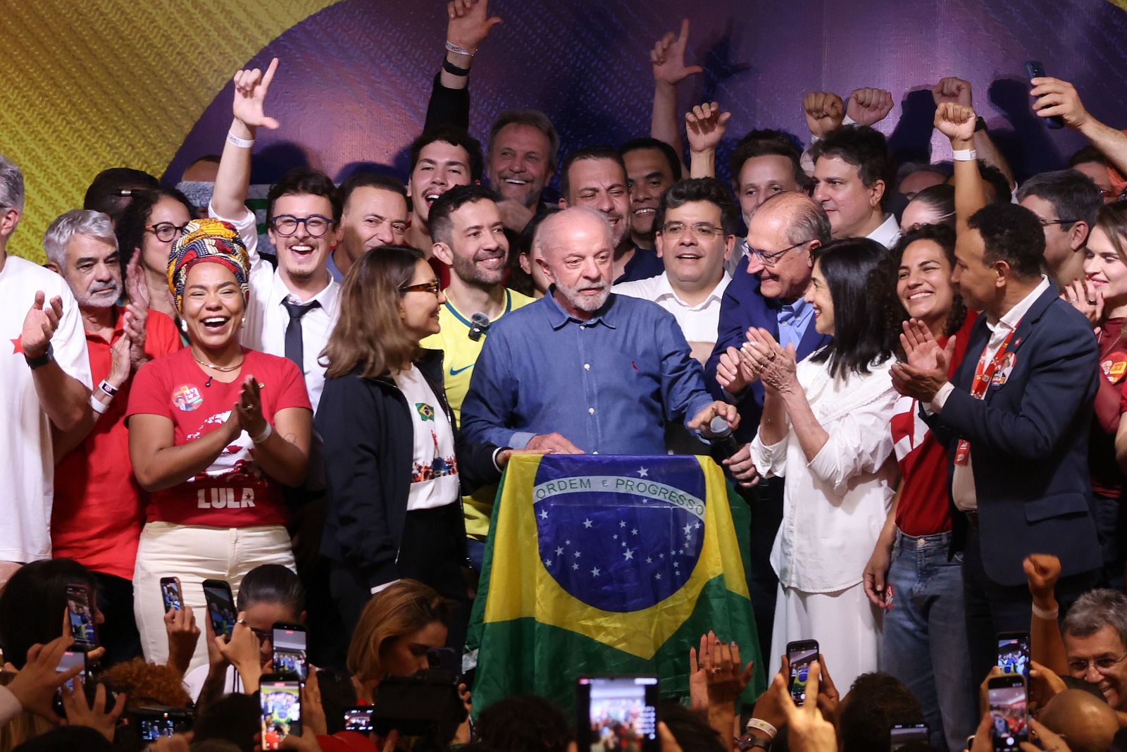 Multitudinaria celebración política con Lula da Silva hablando ante un atril con la bandera de Brasil, rodeado por colaboradores que aplauden y asistentes grabando con móviles.