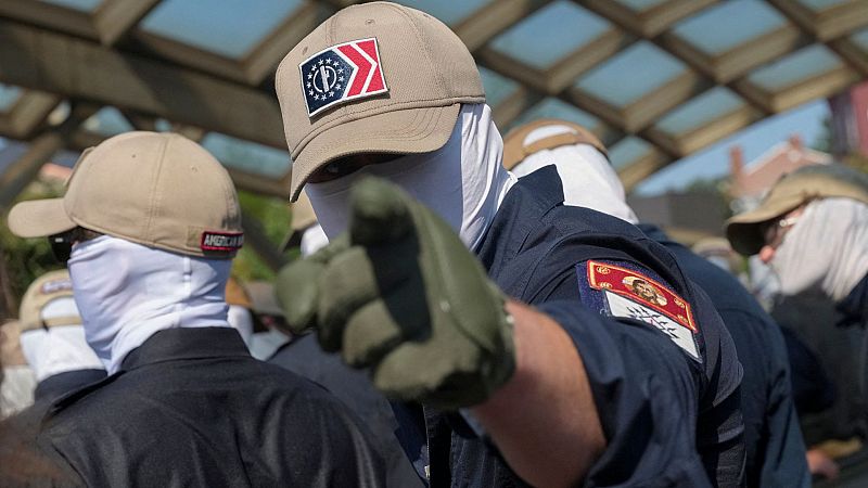 Varios individuos con pasamontañas blancos y gorras caqui ocultan sus rostros, mientras uno de ellos apunta amenazante a la cámara.
