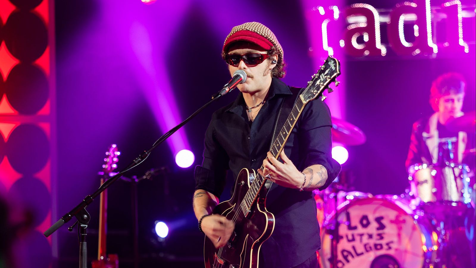 Guitarrista con boina y gafas de sol tocando en directo en un plató iluminado con luces de tonos magenta y púrpura.