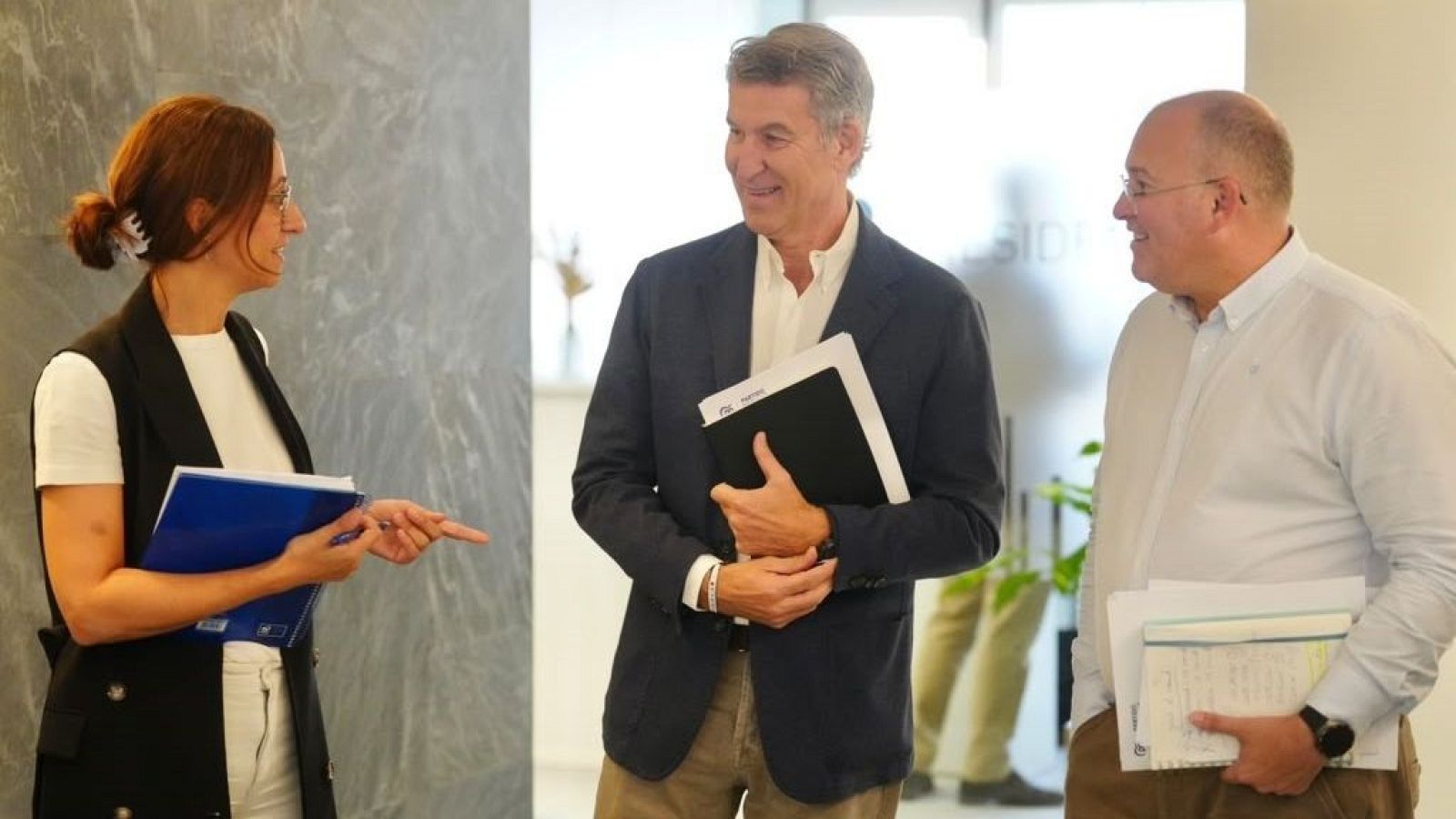 Alberto Núñez Feijóo conversa sonriente en un pasillo junto a Marta Varela y Miguel Tellado, todos con carpetas y documentos de trabajo.