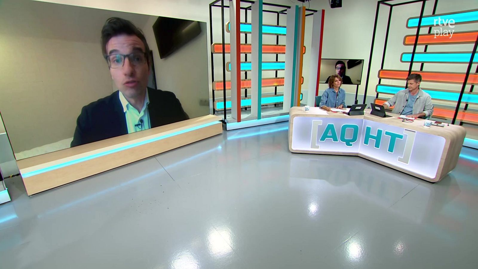 Plató de televisión con dos presentadores sentados tras una mesa blanca iluminada y una pantalla lateral que muestra a un especialista por videollamada.
