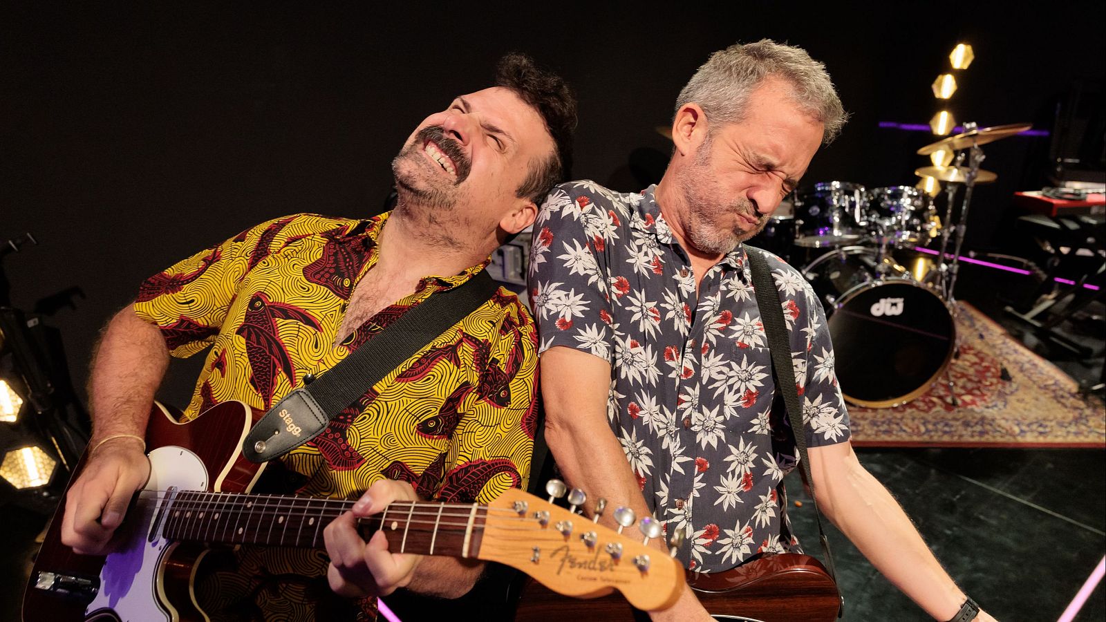 Dos presentadores con camisas estampadas posan como guitarristas de rock en un plató con batería, alfombra y focos.