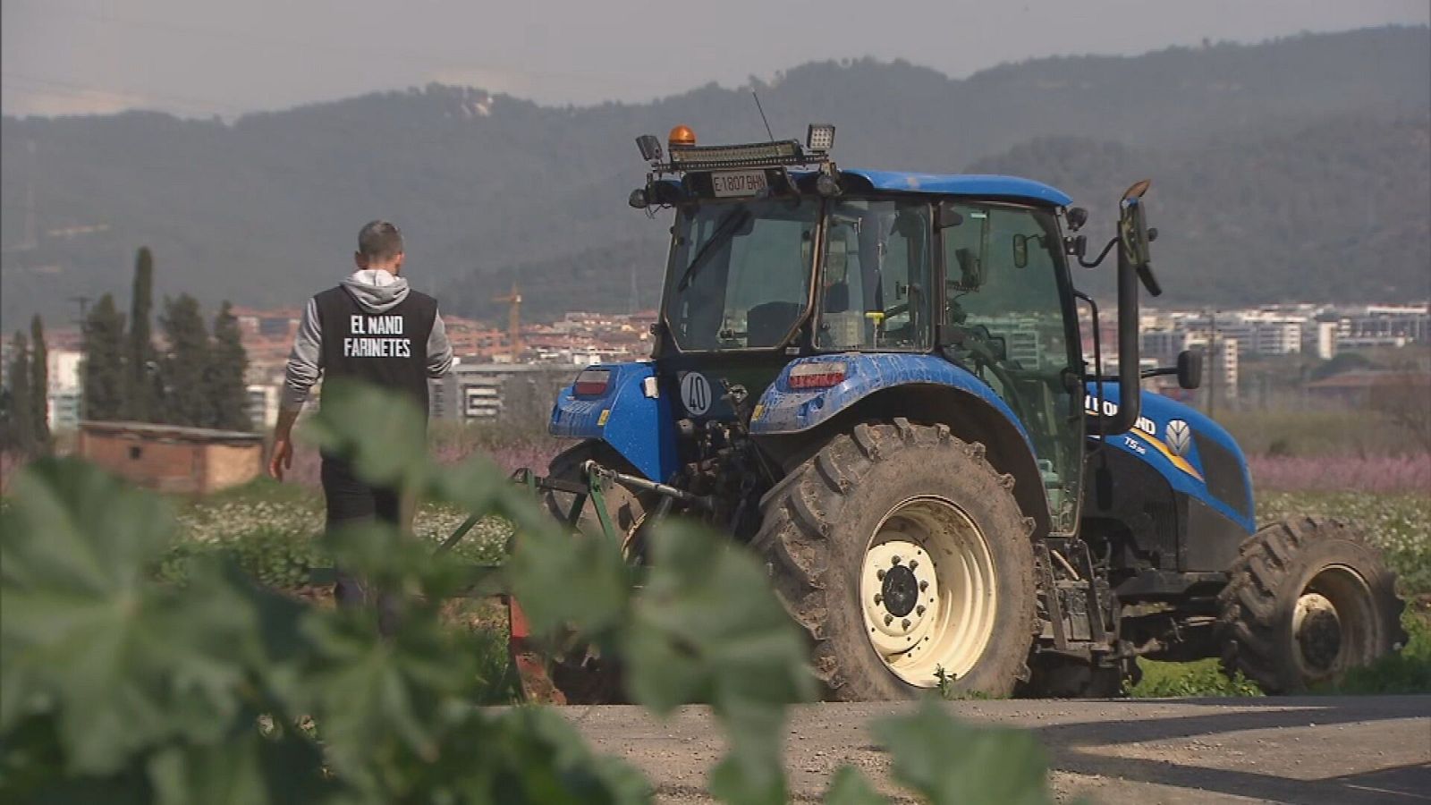 Un tractor azul New Holland y un hombre con chaleco negro caminan por un camino rural con campos de cultivo al fondo.