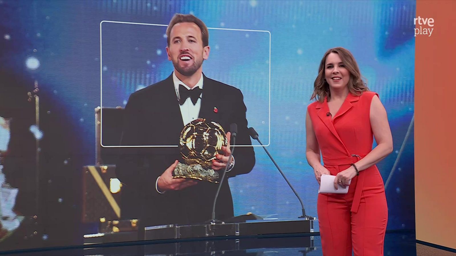 La presentadora Lara Gandarillas, con mono rojo, sonríe en un plató con una pantalla gigante que muestra al futbolista Harry Kane sosteniendo un trofeo.