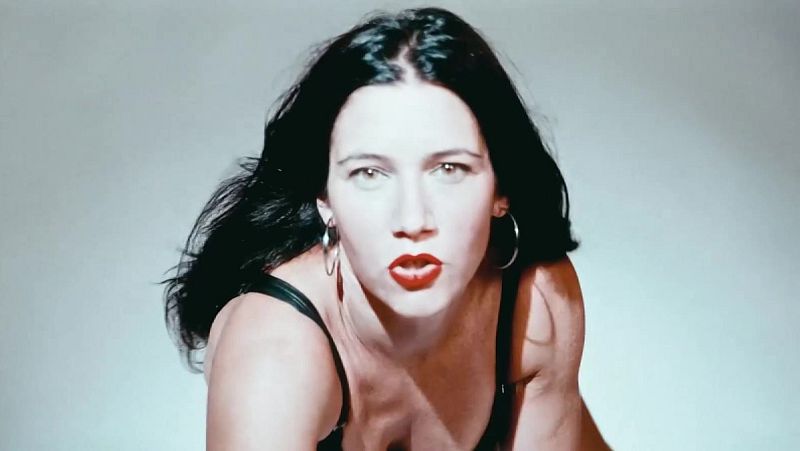 Retrato de Eva Amaral mirando intensamente a la cámara, con melena negra, labios rojos y prenda de tirantes sobre fondo gris.