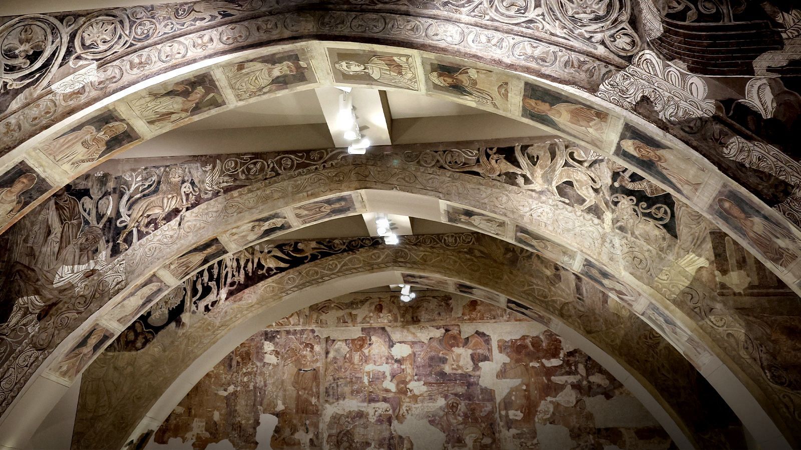 Montaje expositivo de pinturas murales románicas sobre arcos apuntados que recrean la arquitectura original, con frescos de motivos vegetales y personajes bíblicos en tonos pardos y rojizos.