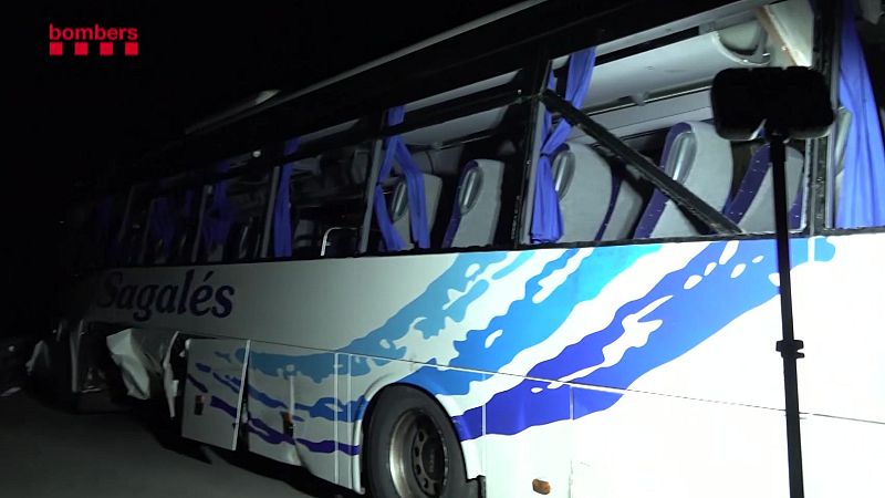 Imagen nocturna del lateral izquierdo de un autobús accidentado con la carrocería dañada y ventanillas rotas, iluminado por un foco de emergencias.