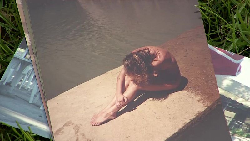 Fotografía artística de una mujer desnuda en postura introspectiva junto al agua, apoyada sobre césped verde.