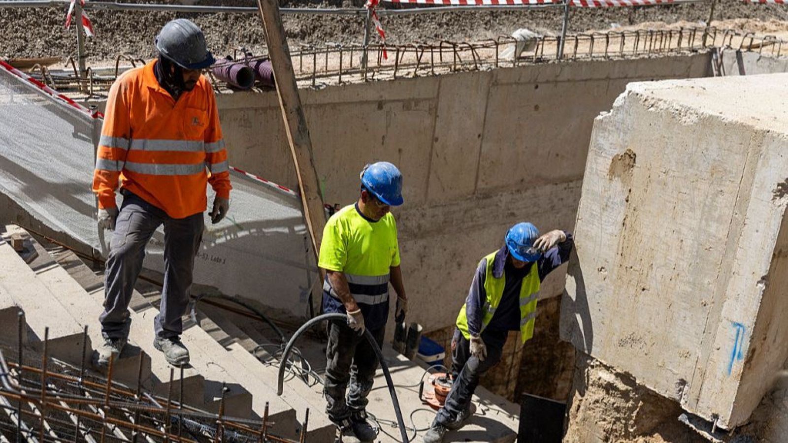 Tres obreros con ropa de alta visibilidad y cascos trabajan en una zanja de construcción bajo el sol.