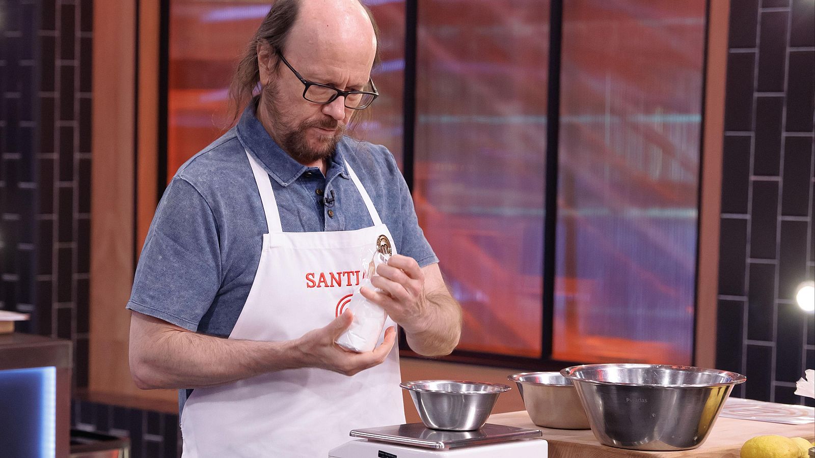 El actor Santiago Segura, con gafas y delantal blanco, cocina concentrado en el plató de MasterChef junto a una báscula y utensilios.