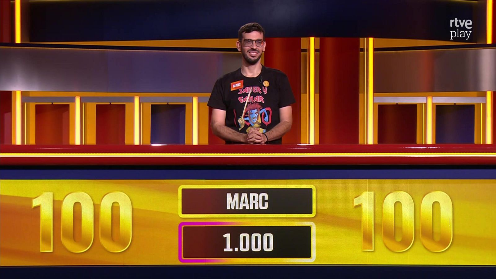 El concursante Marc sonríe en el plató de un concurso televisivo, apoyado en un mostrador que señala la cifra de 1.000 puntos y su programa número 100.