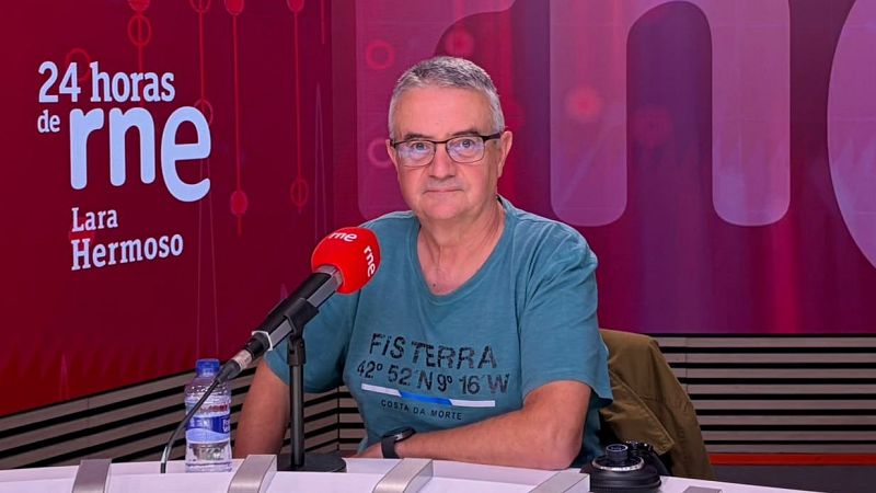 Hombre maduro de pelo canoso y gafas sentado en un estudio de radio ante un micrófono con el logotipo de rne, luciendo una camiseta azul con texto.