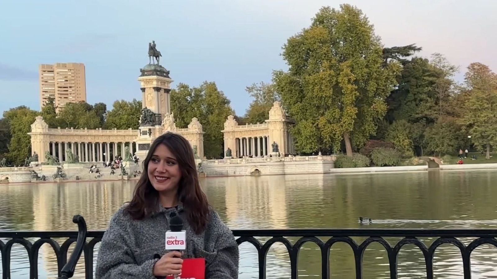 Joven con abrigo gris y micr�fono de radio en el Parque del Buen Retiro, con el estanque y el monumento al fondo.