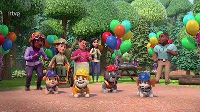 Cachorros constructores de la serie animada 'Equipo Rubble' posan alegres en un claro del bosque decorado con globos y una mesa de pícnic, rodeados por habitantes del pueblo.