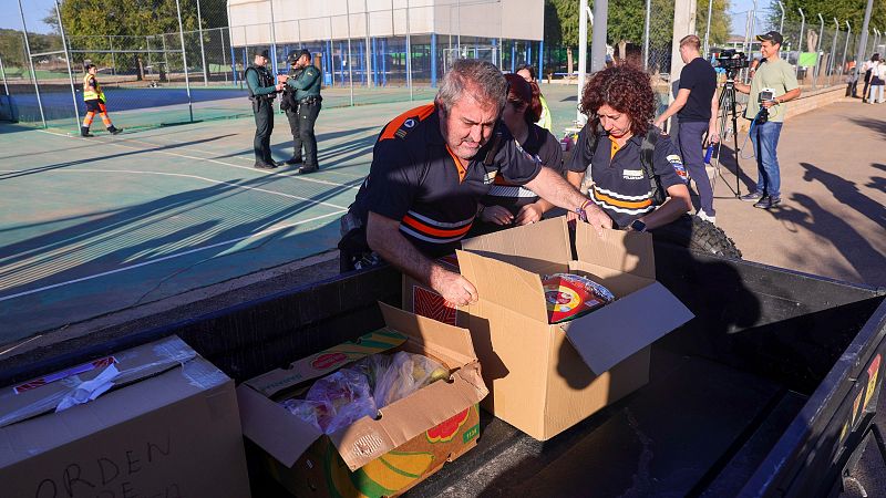 Miembros de Protección Civil descargan cajas con alimentos y suministros desde un vehículo junto a una pista deportiva mientras son grabados por la prensa.