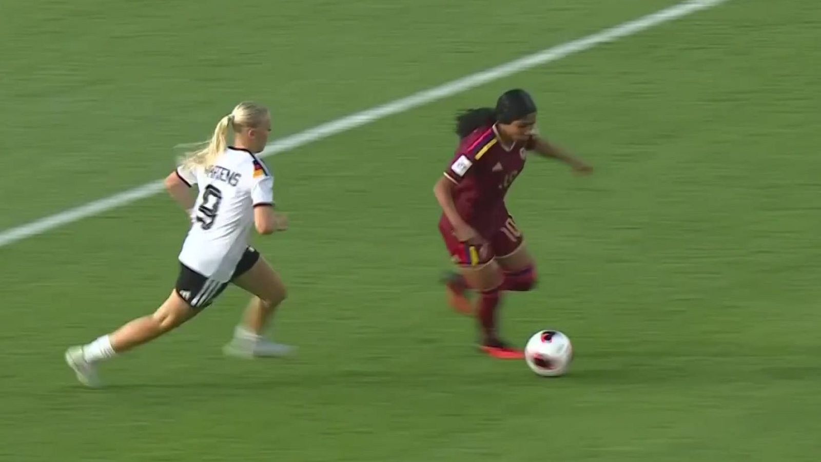 Dos futbolistas disputan un balón en carrera sobre césped verde, con la jugadora colombiana conduciendo la pelota y la alemana persiguiéndola de cerca.