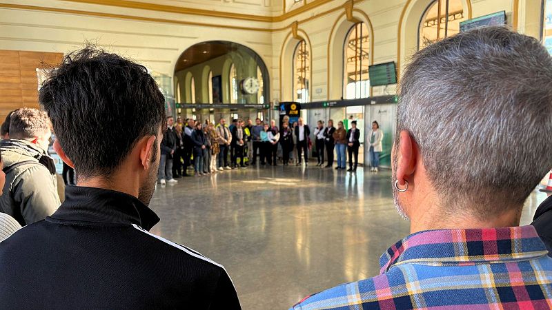 Grupo de personas guardando un minuto de silencio en el vestíbulo de una estación de trenes con arquitectura clásica y suelo de mármol pulido.