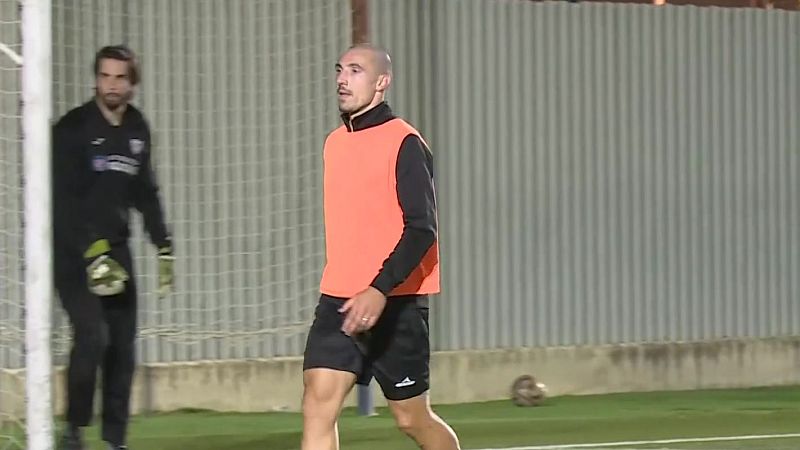 Futbolista con peto naranja y pantalón negro camina por el campo de entrenamiento junto a la portería.