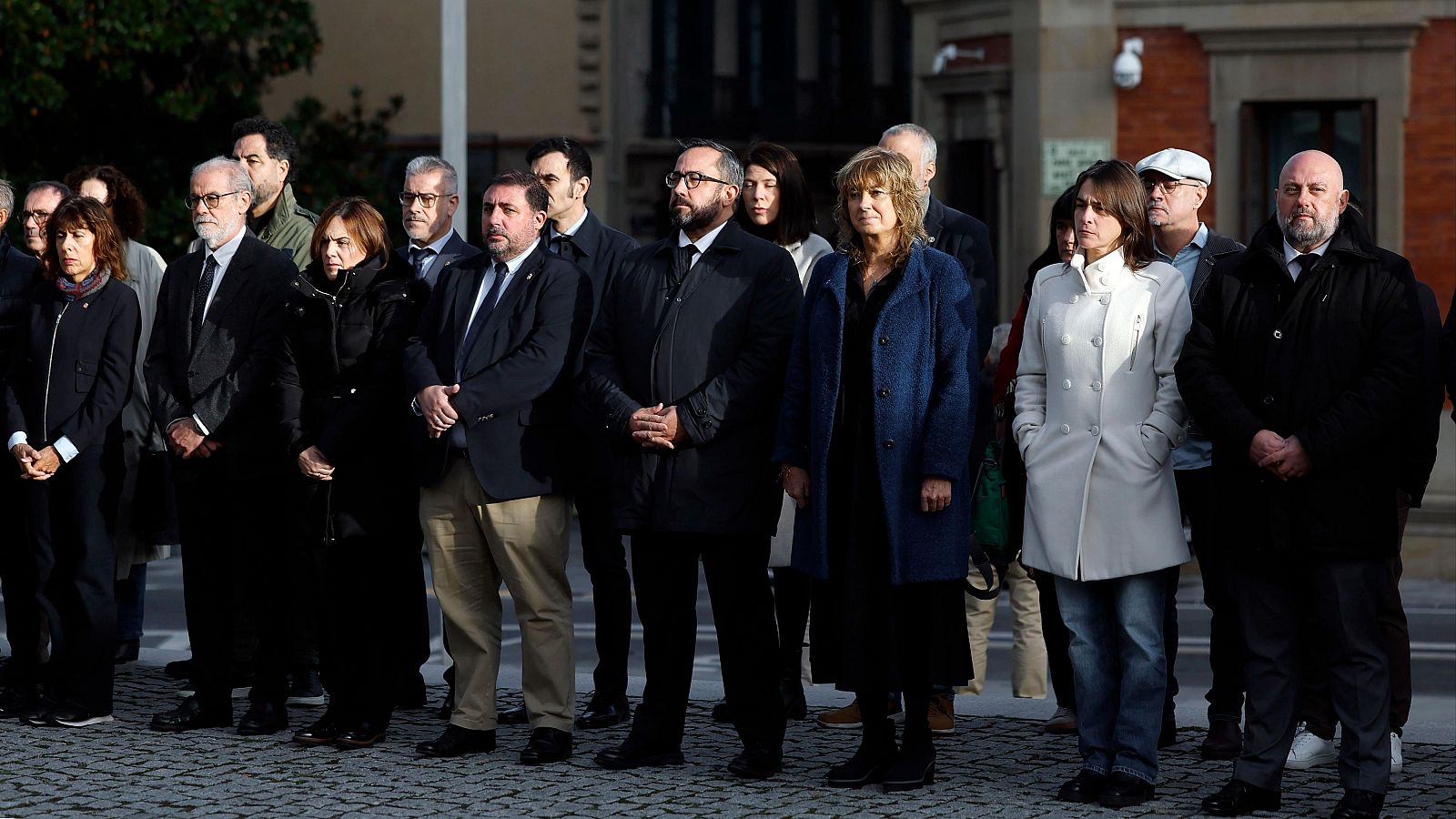 Representantes parlamentarios y del Gobierno foral guardan un solemne minuto de silencio formados en línea sobre un suelo adoquinado.