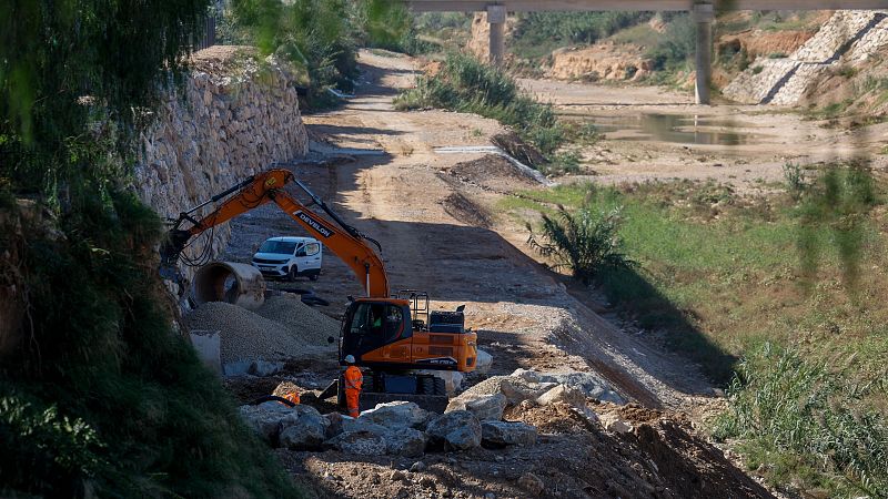 Obras de reconstrucción con una excavadora naranja y un operario en el cauce seco de un barranco, junto a un tubo de hormigón y un muro de contención de piedra.
