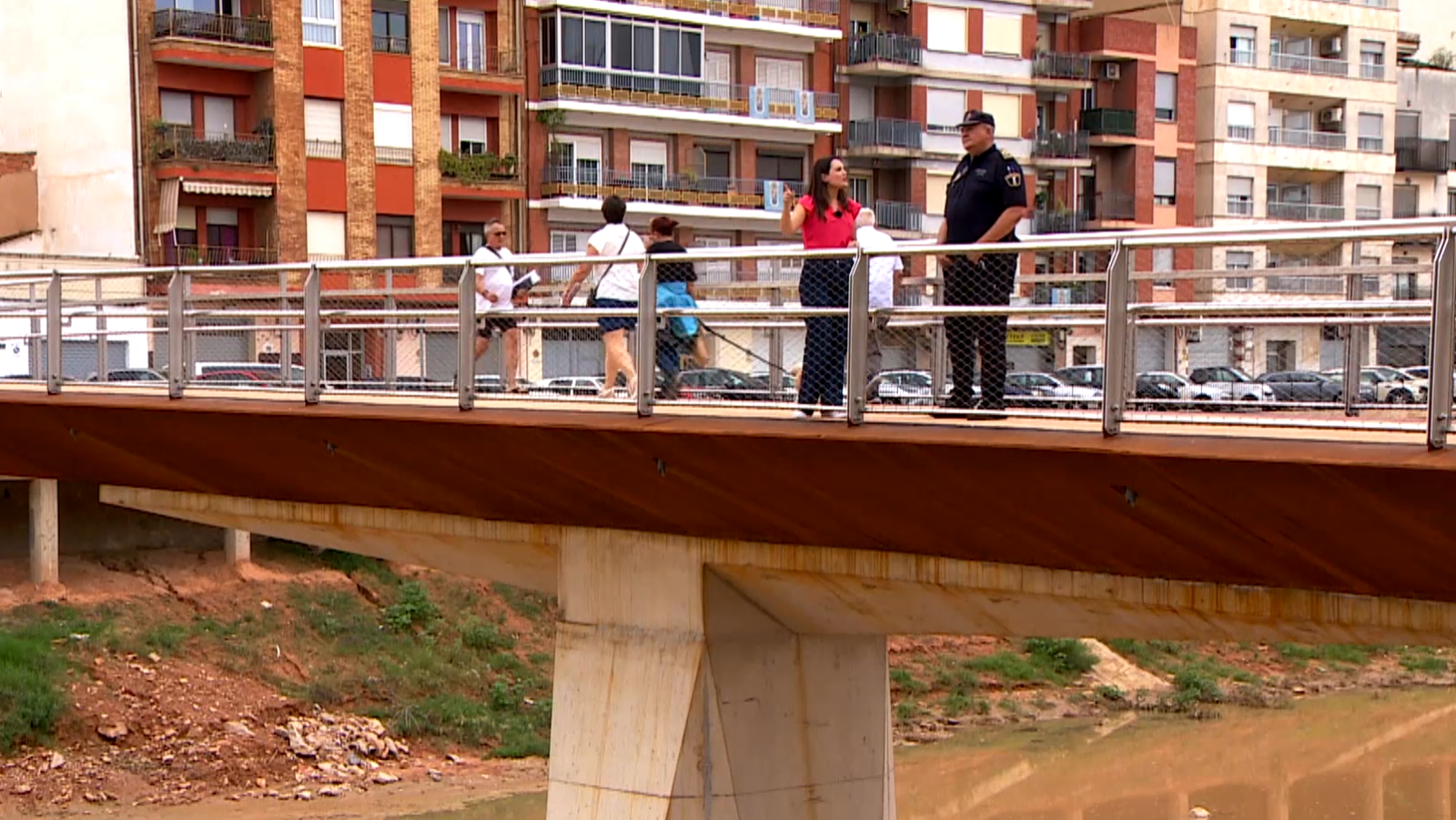 Pasarela peatonal moderna donde una reportera de blusa fucsia conversa con un policía uniformado, con edificios residenciales al fondo.