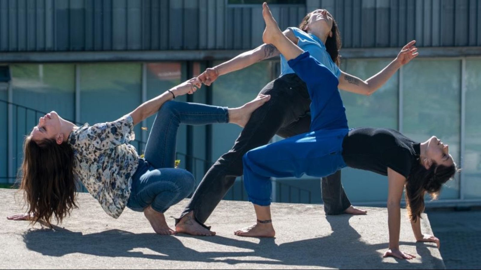 Tres bailarinas descalzas realizan una compleja figura de danza contempor�nea sobre hormig�n, combinando equilibrio, contrapeso y expresividad corporal al aire libre.