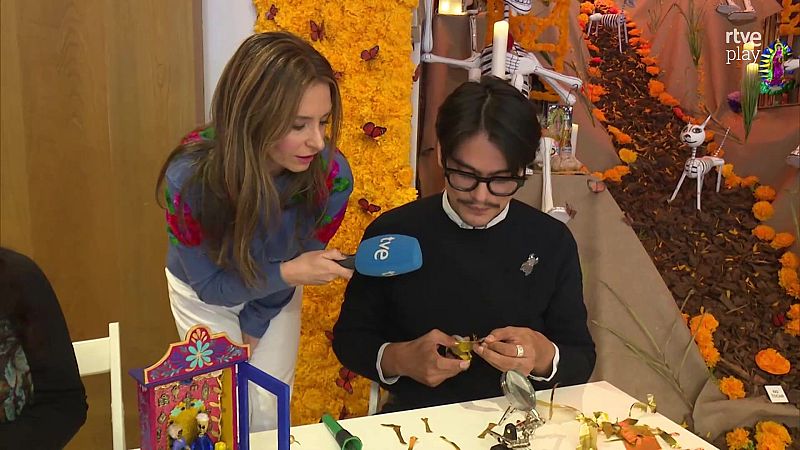 Una reportera entrevista a un artesano mexicano que elabora una miniatura en un espacio decorado por el Día de Muertos.