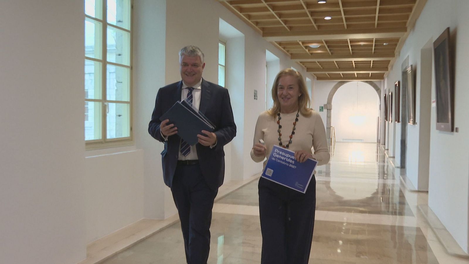 Dos políticos caminan sonrientes por un pasillo luminoso con ventanales y retratos, portando documentos oficiales con el escudo institucional.