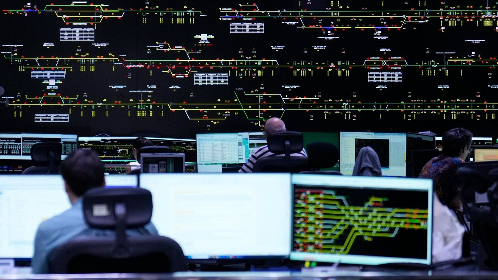 Centro de control ferroviario con operadores supervisando pantallas frente a un gran panel mural interactivo con diagramas de vías y líneas luminosas.