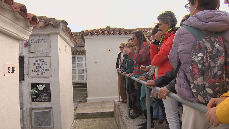 Un grupo de personas asiste a una visita guiada en un cementerio, contemplando una lápida de mármol blanco bajo un cielo nublado.