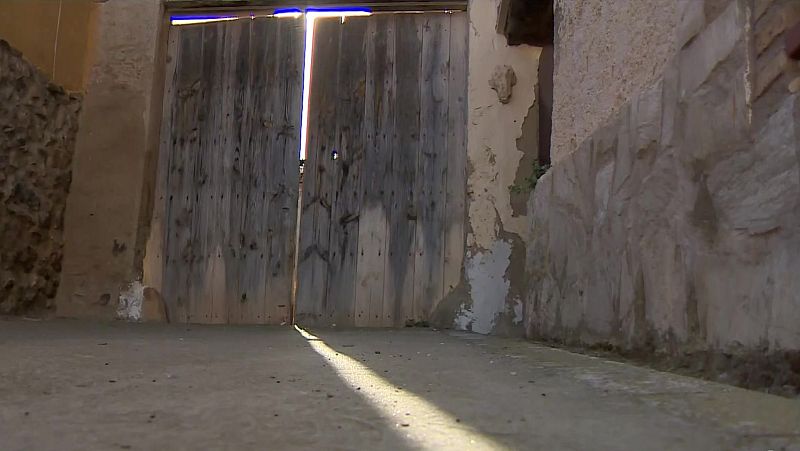 Portón rústico de madera al final de un estrecho paso, donde la luz solar se filtra intensamente entre fachadas de viviendas rurales con muros de piedra y paredes deterioradas.