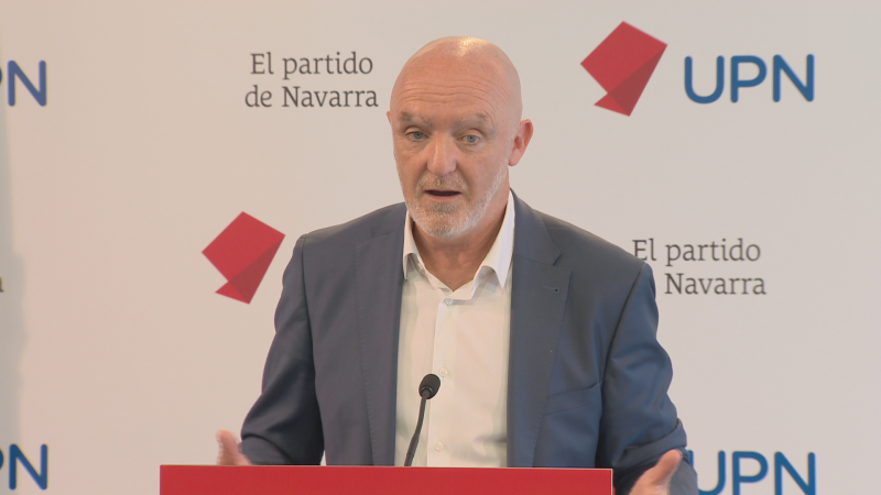 Plano medio de Javier Esparza interviniendo ante un atril rojo con micrófono, vistiendo americana azul y camisa blanca.