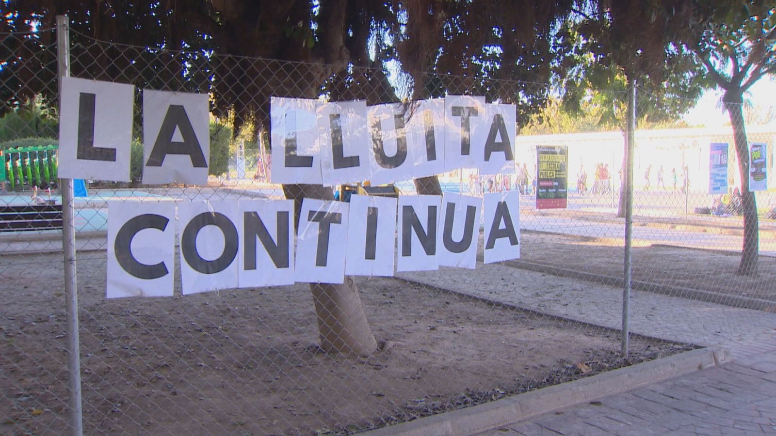 Pancarta improvisada sobre una valla con el lema en valenciano «LA LLUITA CONTINUA», frente a un colegio con aulas prefabricadas.