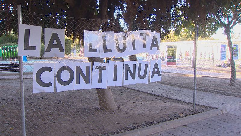 Pancarta improvisada sobre una valla con el lema en valenciano «LA LLUITA CONTINUA», frente a un colegio con aulas prefabricadas.