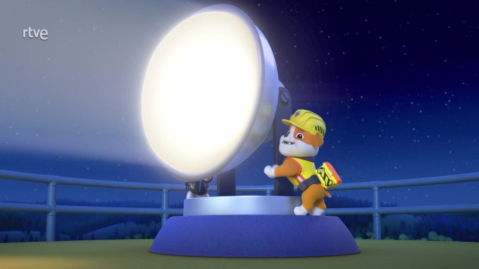 El cachorro Rubble, con casco y chaleco amarillo, ajusta un gran foco reflector luminoso sobre una plataforma nocturna con barandilla azul y cielo estrellado.