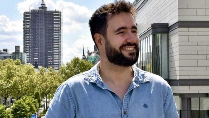Hombre con barba y pelo castaño rizado, sonriendo, vestido con camisa vaquera ante un fondo urbano soleado.