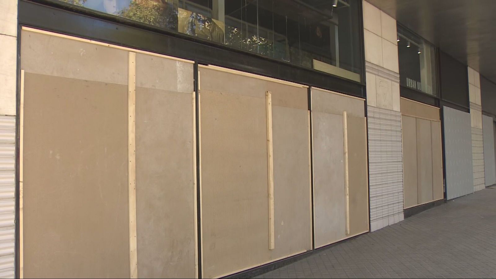 Fachada de tiendas urbanas con los escaparates tapiados con planchas de madera y listones de refuerzo en una acera vacía.