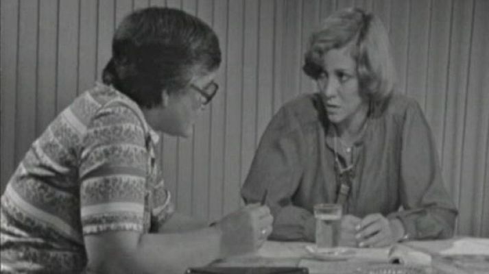 Imagen en blanco y negro de dos personas en una entrevista; una de ellas, Marta Mata, sentada a una mesa con papeles y un vaso.