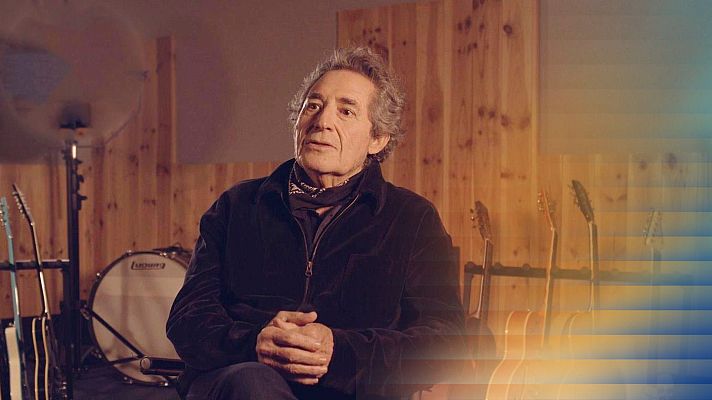 Miguel Ríos, cantante español, en un estudio con guitarras y batería Ludwig. Cabello gris, chaqueta oscura y pañuelo al cuello. Fondo de madera con iluminación cálida.