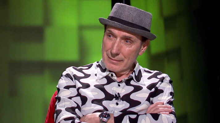 Actor español sentado en un coloquio, con sombrero gris, camisa estampada y reloj. Expresión seria y levemente sonriente.